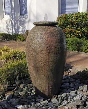 Springbrunnen Krug Antico (110 cm hoch, 60 cm Ø) aus gesandstrahltem Keramik in antiker Optik, mit Wasserstrahl, aufgestellt in einem mediterranen Garten bei Dämmerung mit Beleuchtung.
