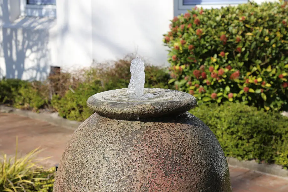 Springbrunnen Krug Antico (110 cm hoch, 60 cm Ø) aus gesandstrahltem Keramik in antiker Optik, mit Wasserstrahl, aufgestellt in einem mediterranen Garten bei Dämmerung mit Beleuchtung.
