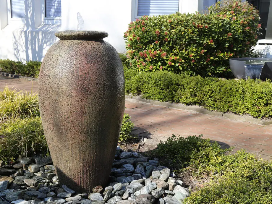 Springbrunnen Krug Antico (110 cm hoch, 60 cm Ø) aus gesandstrahltem Keramik in antiker Optik, mit Wasserstrahl, aufgestellt in einem mediterranen Garten bei Dämmerung mit Beleuchtung.