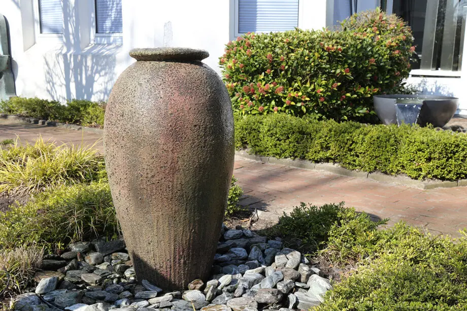 Springbrunnen Krug Antico (110 cm hoch, 60 cm Ø) aus gesandstrahltem Keramik in antiker Optik, mit Wasserstrahl, aufgestellt in einem mediterranen Garten bei Dämmerung mit Beleuchtung.
