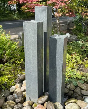 Wasserobjekt Zinkart-Säulen Mod. ZA 1 mit drei Zink-Säulen (110, 95, 80 cm hoch), aufgestellt auf einer Terrasse mit Holzdielen und modernen Pflanzgefäßen.