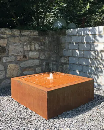 Moderner Cortenstahl-Kubus-Wassertisch als Terrassenbrunnen mit kleiner Wasserfontäne auf Kiesfläche vor Steinmauer.