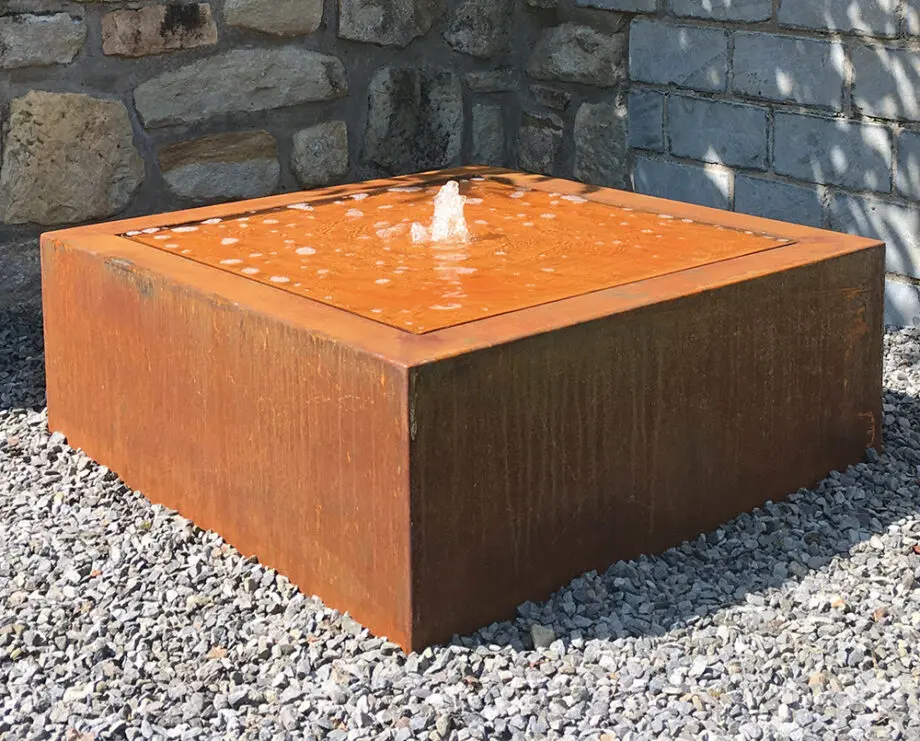 Moderner Cortenstahl-Kubus-Wassertisch als Terrassenbrunnen mit kleiner Wasserfontäne auf Kiesfläche vor Steinmauer.