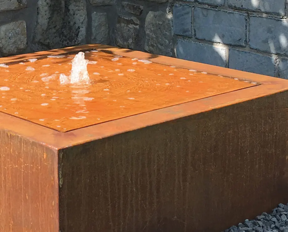 Moderner Cortenstahl-Kubus-Wassertisch als Terrassenbrunnen mit kleiner Wasserfontäne auf Kiesfläche vor Steinmauer.