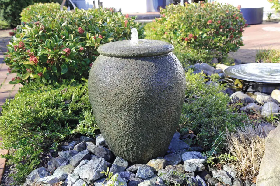 Palermo Keramik-Springbrunnen in antiker Krugform mit sprudelndem Wasser, umgeben von grünen Pflanzen und Steinen in einem Garten.