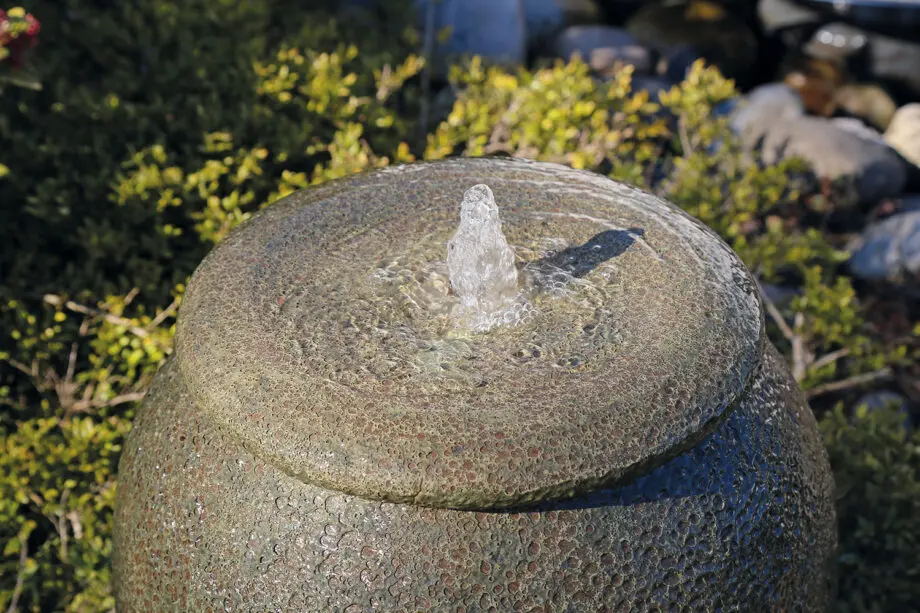 Palermo Keramik-Springbrunnen in antiker Krugform mit sprudelndem Wasser, umgeben von grünen Pflanzen und Steinen in einem Garten.