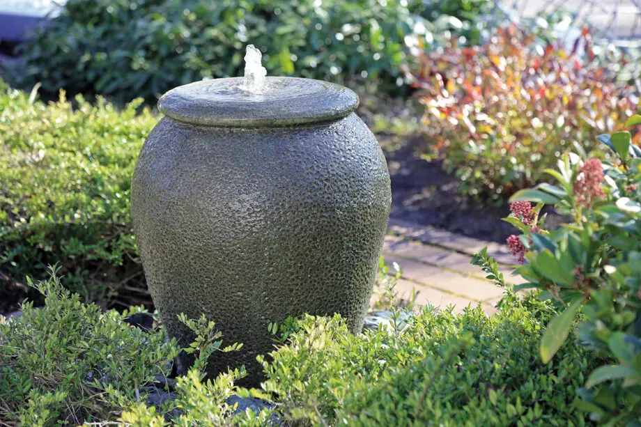 Palermo Keramik-Springbrunnen in antiker Krugform mit sprudelndem Wasser, umgeben von grünen Pflanzen und Steinen in einem Garten.