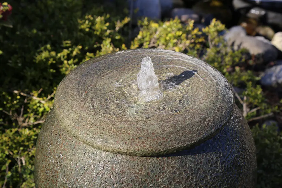 Palermo Keramik-Springbrunnen in antiker Krugform mit sprudelndem Wasser, umgeben von grünen Pflanzen und Steinen in einem Garten.