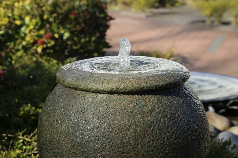 Palermo Keramik-Springbrunnen in antiker Krugform mit sprudelndem Wasser, umgeben von grünen Pflanzen und Steinen in einem Garten.