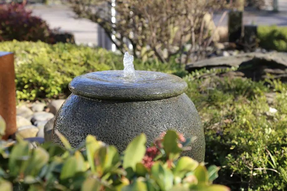 Palermo Keramik-Springbrunnen in antiker Krugform mit sprudelndem Wasser, umgeben von grünen Pflanzen und Steinen in einem Garten.