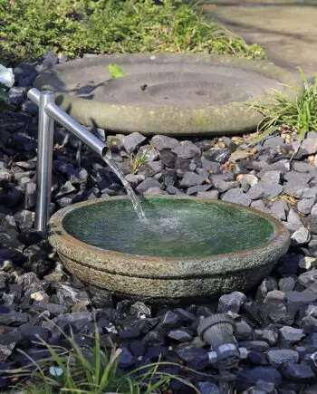 Noto Keramikbrunnen mit schimmernd grüner Wasserschale und Edelstahl-Wasserauslauf, Wasser fließt in die Schale, umgeben von dunklen Steinen in einem Garten.