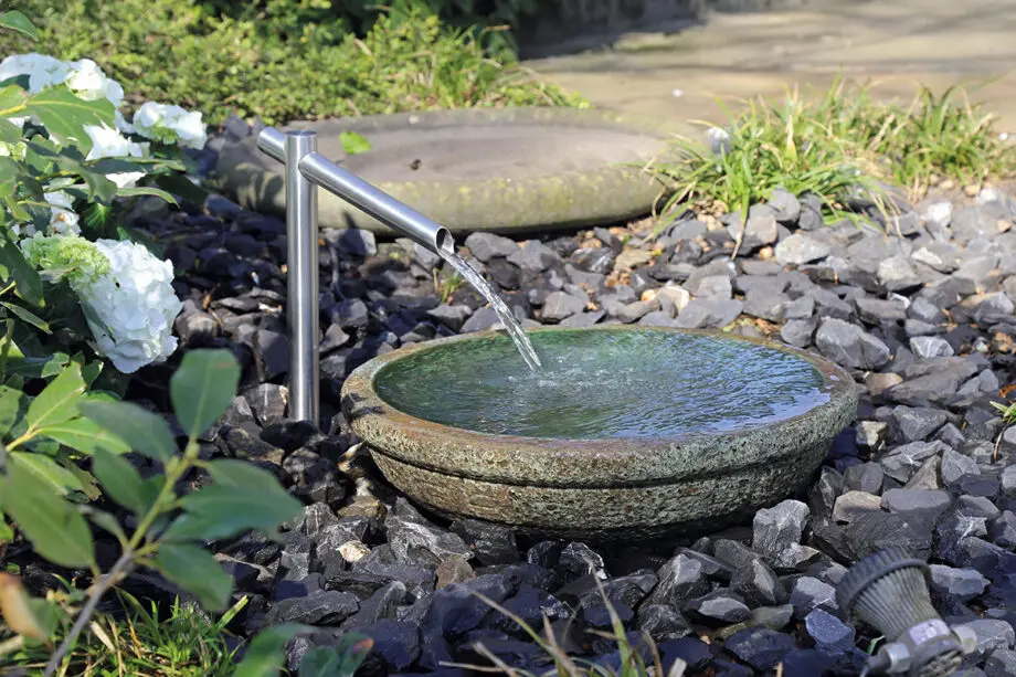 Noto Keramikbrunnen mit schimmernd grüner Wasserschale und Edelstahl-Wasserauslauf, Wasser fließt in die Schale, umgeben von dunklen Steinen in einem Garten.