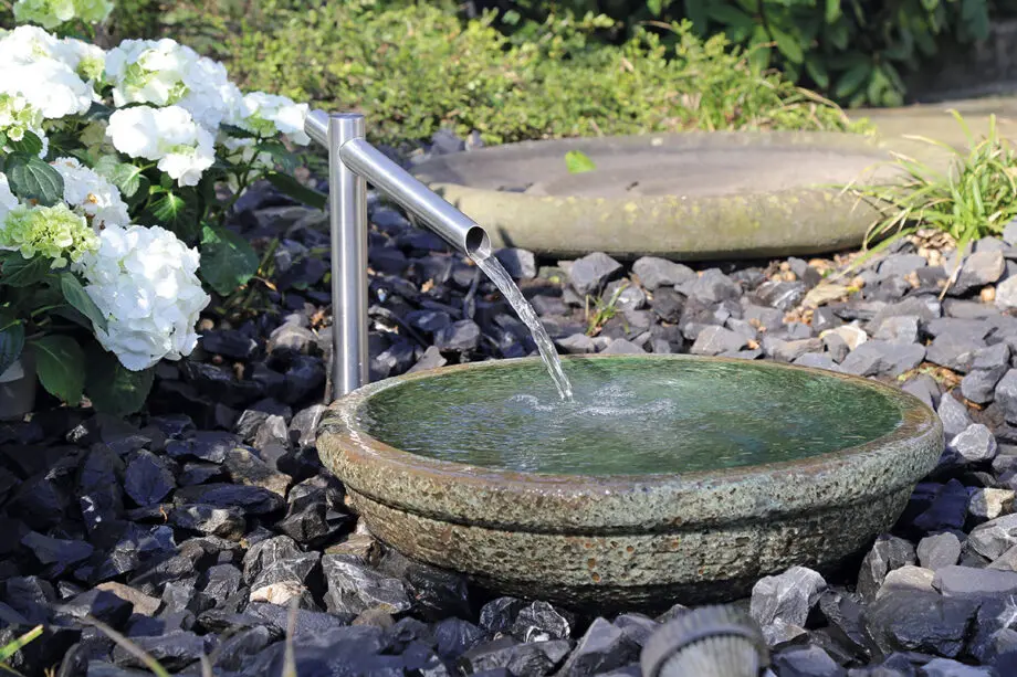 Noto Keramikbrunnen mit schimmernd grüner Wasserschale und Edelstahl-Wasserauslauf, Wasser fließt in die Schale, umgeben von dunklen Steinen in einem Garten.