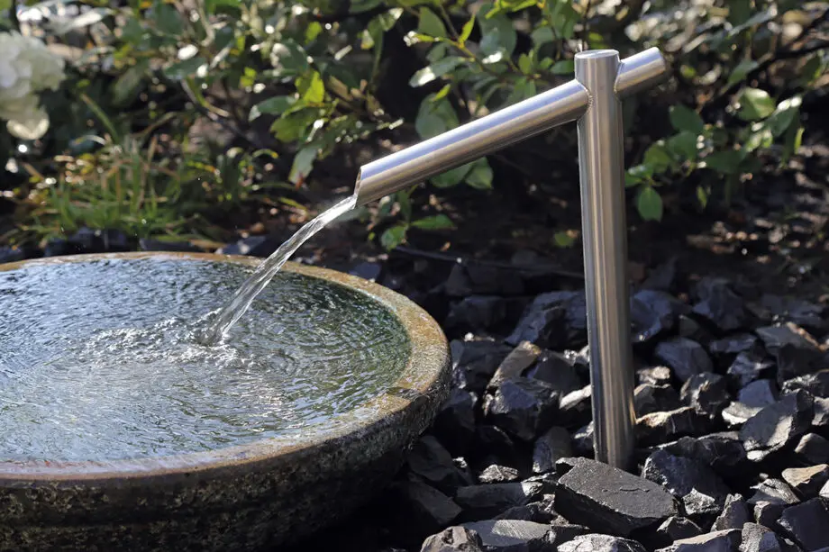 Noto Keramikbrunnen mit schimmernd grüner Wasserschale und Edelstahl-Wasserauslauf, Wasser fließt in die Schale, umgeben von dunklen Steinen in einem Garten.