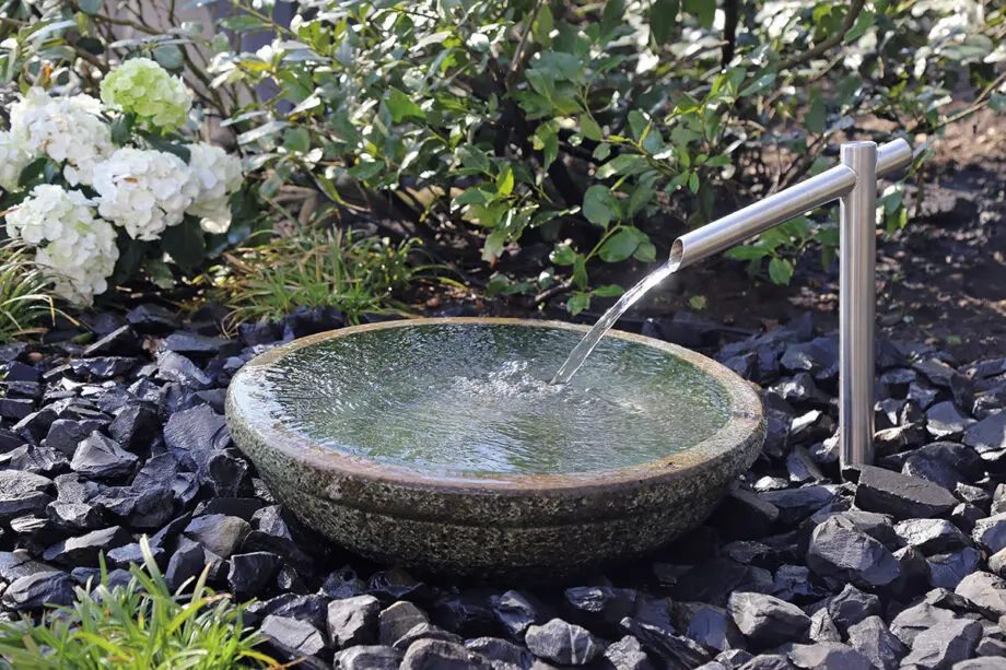 Noto Keramikbrunnen mit schimmernd grüner Wasserschale und Edelstahl-Wasserauslauf, Wasser fließt in die Schale, umgeben von dunklen Steinen in einem Garten.