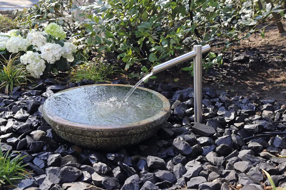 Noto Keramikbrunnen mit schimmernd grüner Wasserschale und Edelstahl-Wasserauslauf, Wasser fließt in die Schale, umgeben von dunklen Steinen in einem Garten.