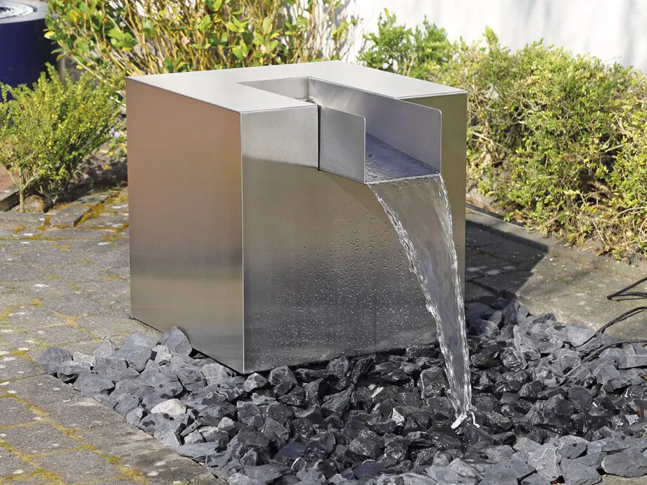Edelstahl Quader Kjaer Designer-Gartenbrunnen mit rechteckigem Wasserauslauf, Wasser fließt in ein Bett aus dunklen Steinen, umgeben von grünen Pflanzen.