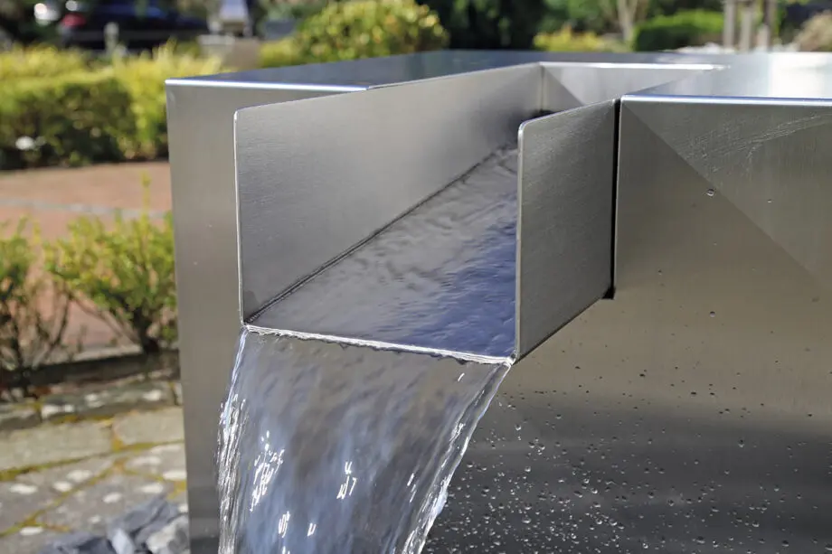 Edelstahl Quader Kjaer Designer-Gartenbrunnen mit rechteckigem Wasserauslauf, Wasser fließt in ein Bett aus dunklen Steinen, umgeben von grünen Pflanzen.