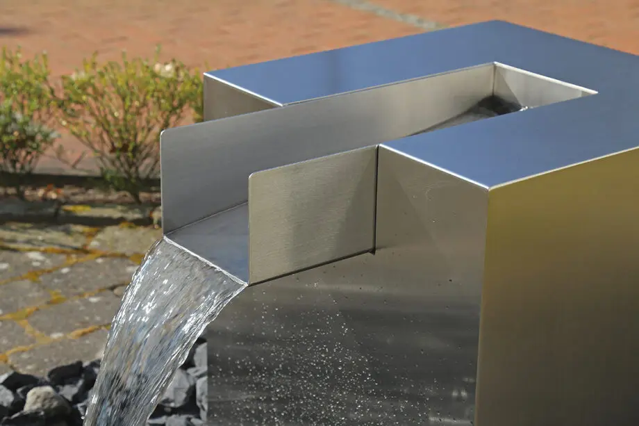 Edelstahl Quader Kjaer Designer-Gartenbrunnen mit rechteckigem Wasserauslauf, Wasser fließt in ein Bett aus dunklen Steinen, umgeben von grünen Pflanzen.