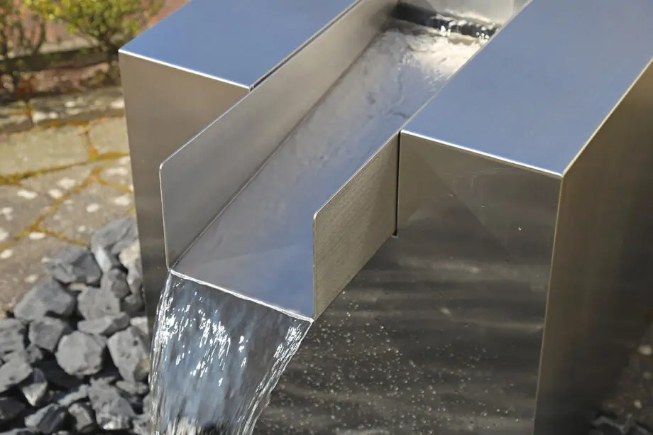 Edelstahl Quader Kjaer Designer-Gartenbrunnen mit rechteckigem Wasserauslauf, Wasser fließt in ein Bett aus dunklen Steinen, umgeben von grünen Pflanzen.
