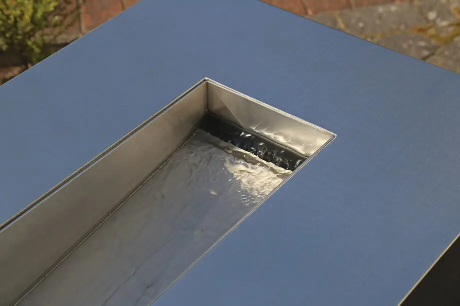 Edelstahl Quader Kjaer Designer-Gartenbrunnen mit rechteckigem Wasserauslauf, Wasser fließt in ein Bett aus dunklen Steinen, umgeben von grünen Pflanzen.