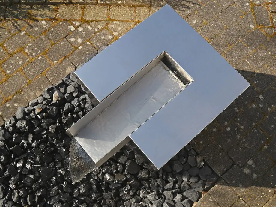 Edelstahl Quader Kjaer Designer-Gartenbrunnen mit rechteckigem Wasserauslauf, Wasser fließt in ein Bett aus dunklen Steinen, umgeben von grünen Pflanzen.
