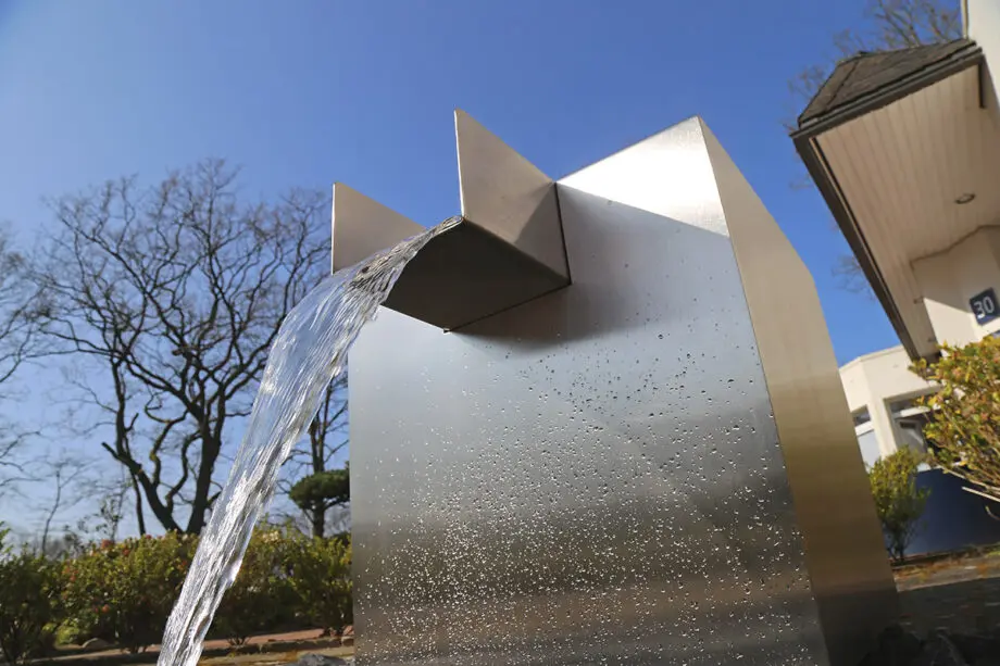 Edelstahl Quader Kjaer Designer-Gartenbrunnen mit rechteckigem Wasserauslauf, Wasser fließt in ein Bett aus dunklen Steinen, umgeben von grünen Pflanzen.