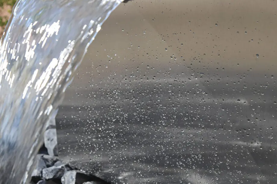 Edelstahl Quader Kjaer Designer-Gartenbrunnen mit rechteckigem Wasserauslauf, Wasser fließt in ein Bett aus dunklen Steinen, umgeben von grünen Pflanzen.