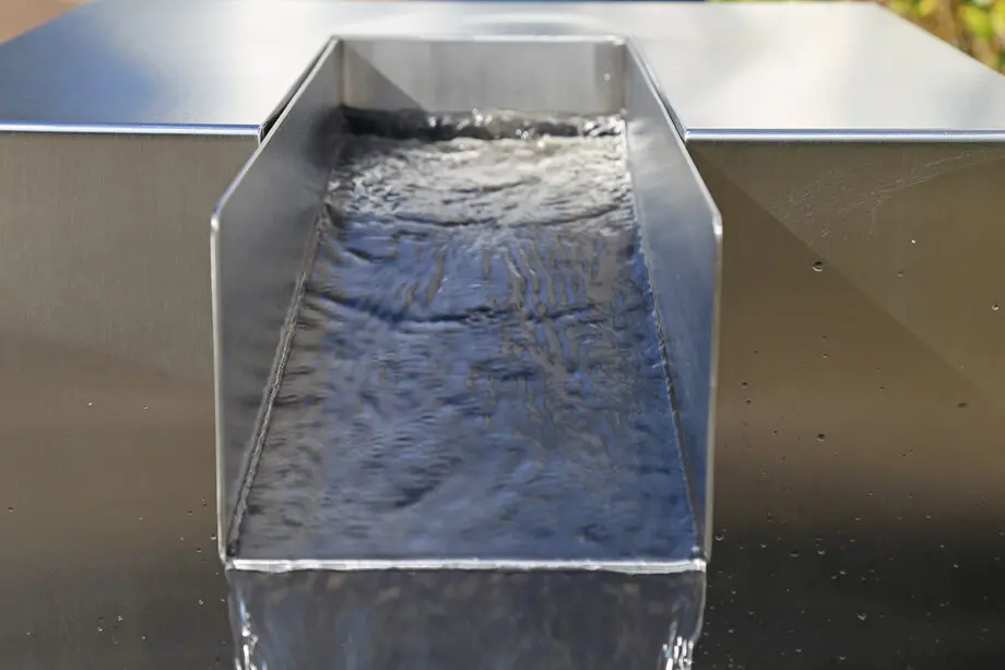 Edelstahl Quader Kjaer Designer-Gartenbrunnen mit rechteckigem Wasserauslauf, Wasser fließt in ein Bett aus dunklen Steinen, umgeben von grünen Pflanzen.