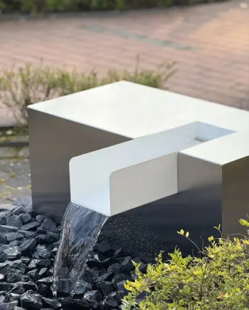 Edelstahl Kubus Kjaer Designer-Gartenbrunnen mit rechteckigem Wasserauslauf, Wasser fließt in ein offenes Wasserbecken, umgeben von modernen Gartenpflanzen.