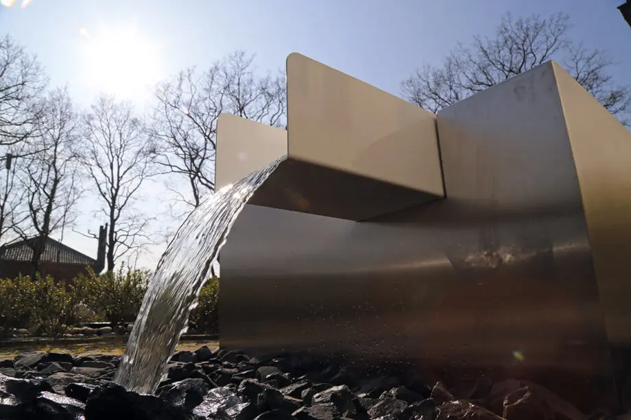 Edelstahl Kubus Kjaer Designer-Gartenbrunnen mit rechteckigem Wasserauslauf, Wasser fließt in ein offenes Wasserbecken, umgeben von modernen Gartenpflanzen.