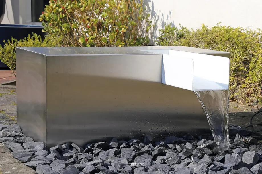 Edelstahl Kubus Kjaer Designer-Gartenbrunnen mit rechteckigem Wasserauslauf, Wasser fließt in ein offenes Wasserbecken, umgeben von modernen Gartenpflanzen.