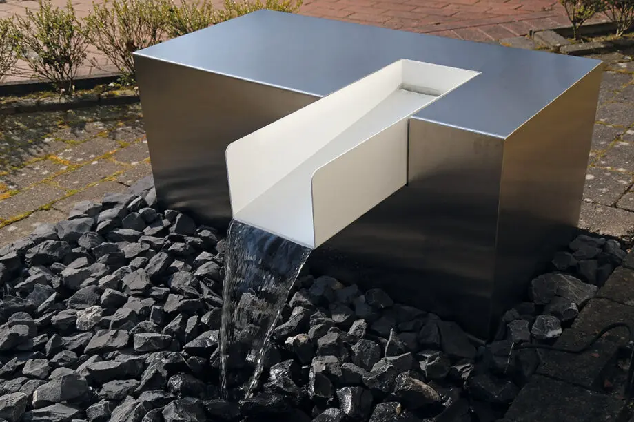 Edelstahl Kubus Kjaer Designer-Gartenbrunnen mit rechteckigem Wasserauslauf, Wasser fließt in ein offenes Wasserbecken, umgeben von modernen Gartenpflanzen.