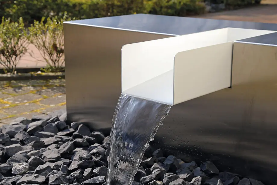 Edelstahl Kubus Kjaer Designer-Gartenbrunnen mit rechteckigem Wasserauslauf, Wasser fließt in ein offenes Wasserbecken, umgeben von modernen Gartenpflanzen.