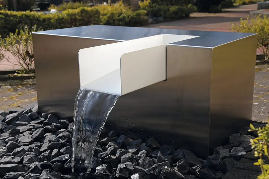 Edelstahl Kubus Kjaer Designer-Gartenbrunnen mit rechteckigem Wasserauslauf, Wasser fließt in ein offenes Wasserbecken, umgeben von modernen Gartenpflanzen.
