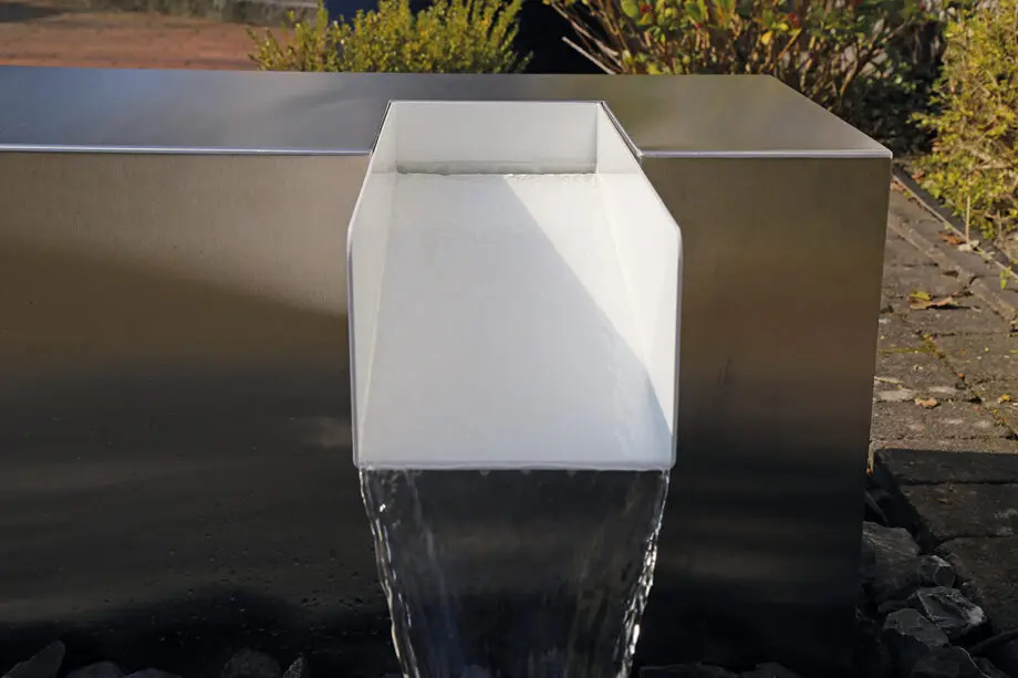 Edelstahl Kubus Kjaer Designer-Gartenbrunnen mit rechteckigem Wasserauslauf, Wasser fließt in ein offenes Wasserbecken, umgeben von modernen Gartenpflanzen.