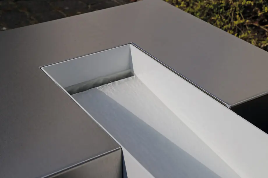 Edelstahl Kubus Kjaer Designer-Gartenbrunnen mit rechteckigem Wasserauslauf, Wasser fließt in ein offenes Wasserbecken, umgeben von modernen Gartenpflanzen.