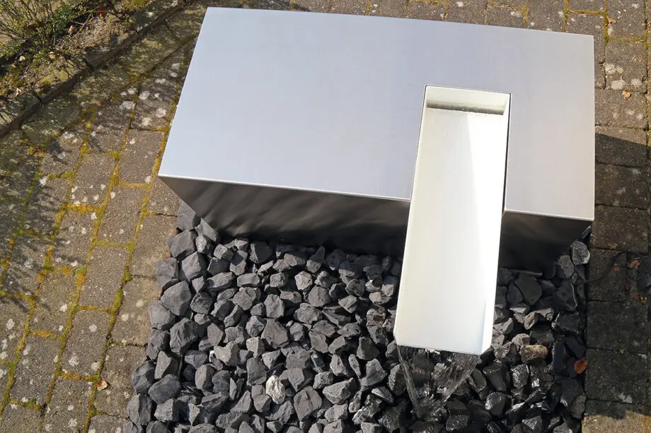 Edelstahl Kubus Kjaer Designer-Gartenbrunnen mit rechteckigem Wasserauslauf, Wasser fließt in ein offenes Wasserbecken, umgeben von modernen Gartenpflanzen.