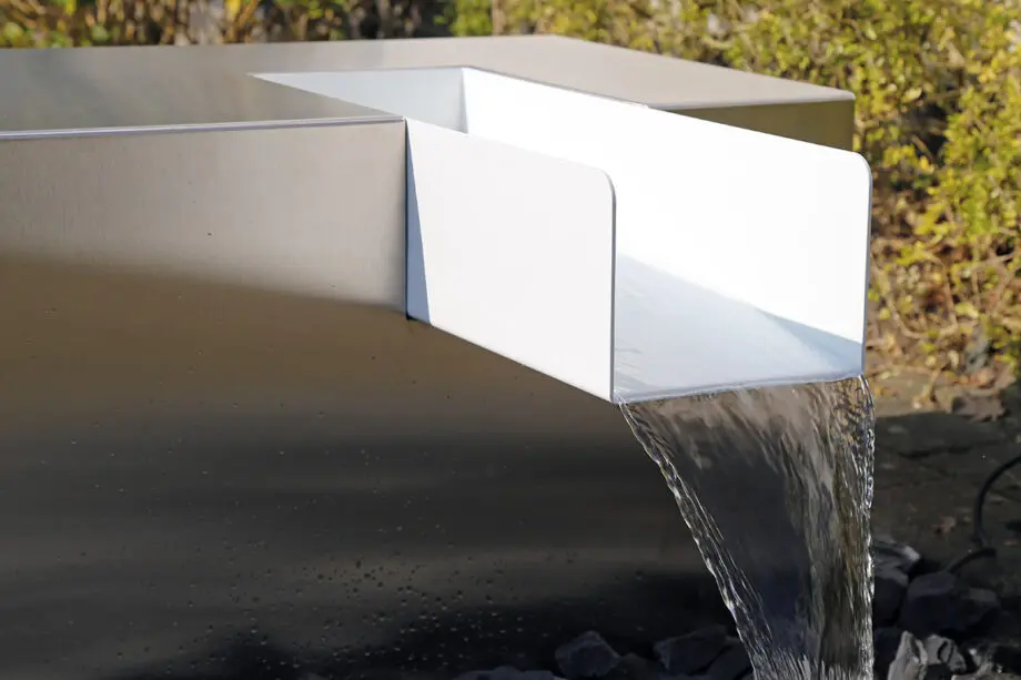 Edelstahl Kubus Kjaer Designer-Gartenbrunnen mit rechteckigem Wasserauslauf, Wasser fließt in ein offenes Wasserbecken, umgeben von modernen Gartenpflanzen.