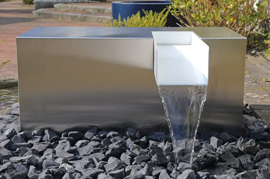 Edelstahl Kubus Kjaer Designer-Gartenbrunnen mit rechteckigem Wasserauslauf, Wasser fließt in ein offenes Wasserbecken, umgeben von modernen Gartenpflanzen.