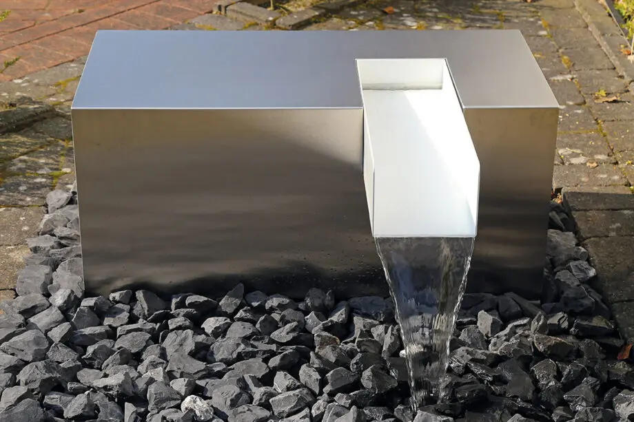 Edelstahl Kubus Kjaer Designer-Gartenbrunnen mit rechteckigem Wasserauslauf, Wasser fließt in ein offenes Wasserbecken, umgeben von modernen Gartenpflanzen.