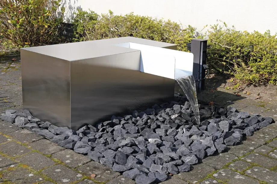 Edelstahl Kubus Kjaer Designer-Gartenbrunnen mit rechteckigem Wasserauslauf, Wasser fließt in ein offenes Wasserbecken, umgeben von modernen Gartenpflanzen.