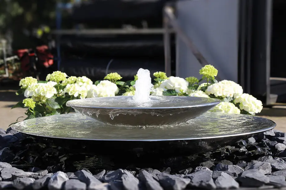 Edelstahl-Schale Duo mit zwei Wasserschalen (100 cm und 60 cm Ø), Sprudelquell in der oberen Schale, aufgestellt in einem modernen Garten mit Kies und Pflanzen.