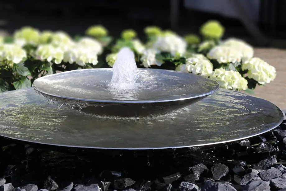 Edelstahl-Schale Duo mit zwei Wasserschalen (100 cm und 60 cm Ø), Sprudelquell in der oberen Schale, aufgestellt in einem modernen Garten mit Kies und Pflanzen.