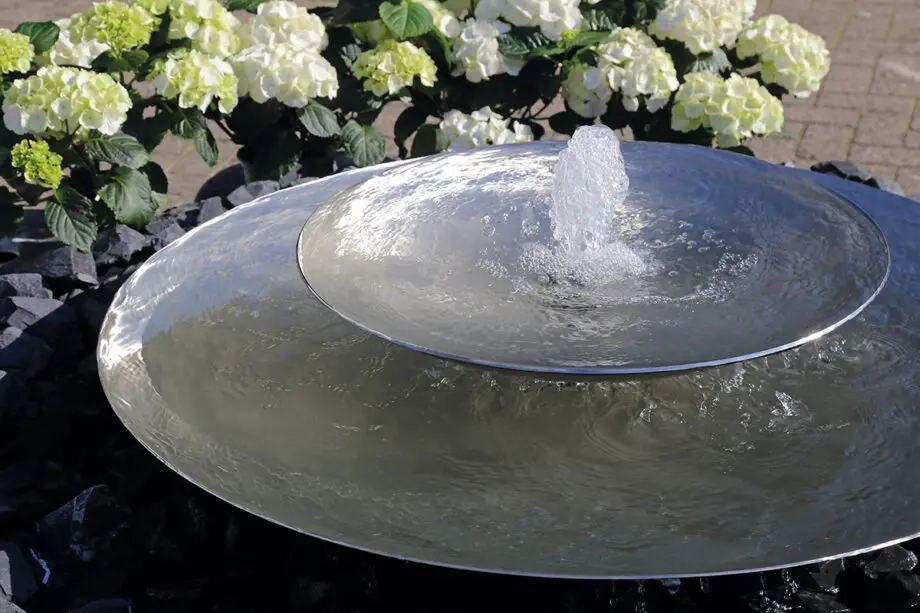 Edelstahl-Schale Duo mit zwei Wasserschalen (100 cm und 60 cm Ø), Sprudelquell in der oberen Schale, aufgestellt in einem modernen Garten mit Kies und Pflanzen.