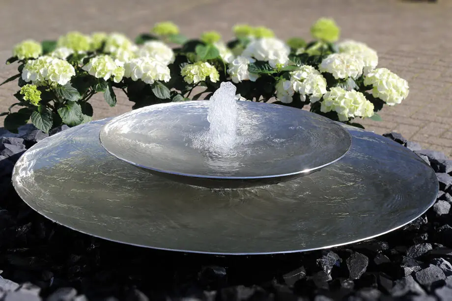 Edelstahl-Schale Duo mit zwei Wasserschalen (100 cm und 60 cm Ø), Sprudelquell in der oberen Schale, aufgestellt in einem modernen Garten mit Kies und Pflanzen.