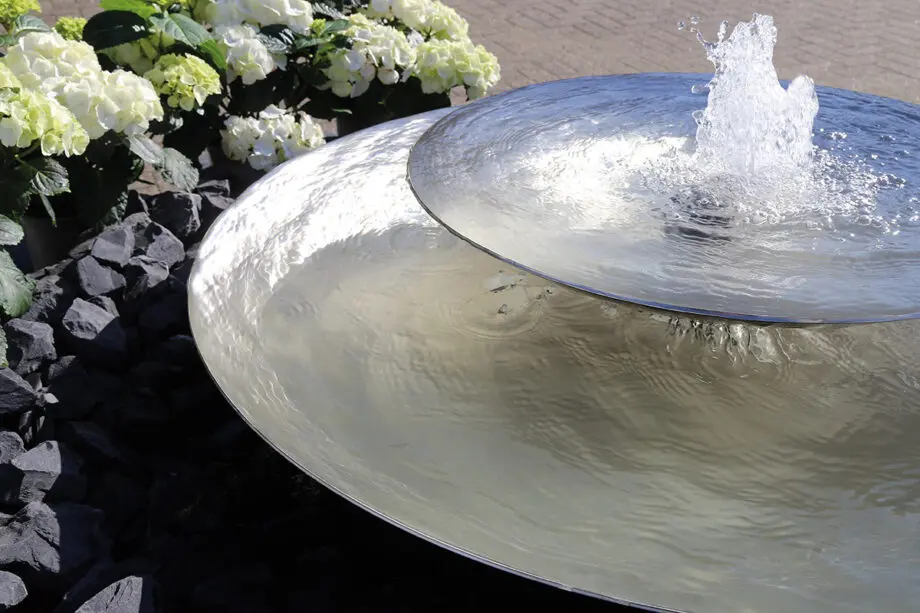Edelstahl-Schale Duo mit zwei Wasserschalen (100 cm und 60 cm Ø), Sprudelquell in der oberen Schale, aufgestellt in einem modernen Garten mit Kies und Pflanzen.