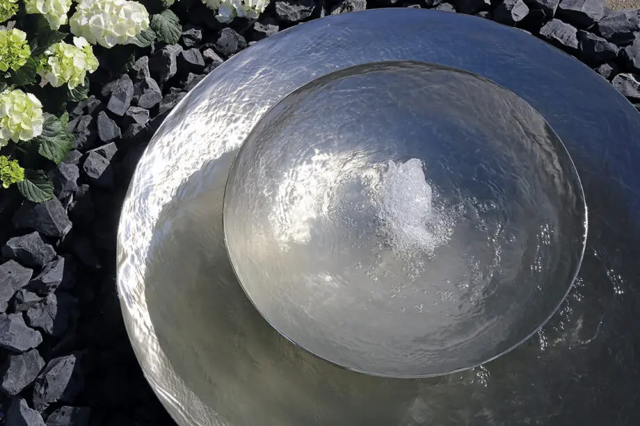 Edelstahl-Schale Duo mit zwei Wasserschalen (100 cm und 60 cm Ø), Sprudelquell in der oberen Schale, aufgestellt in einem modernen Garten mit Kies und Pflanzen.