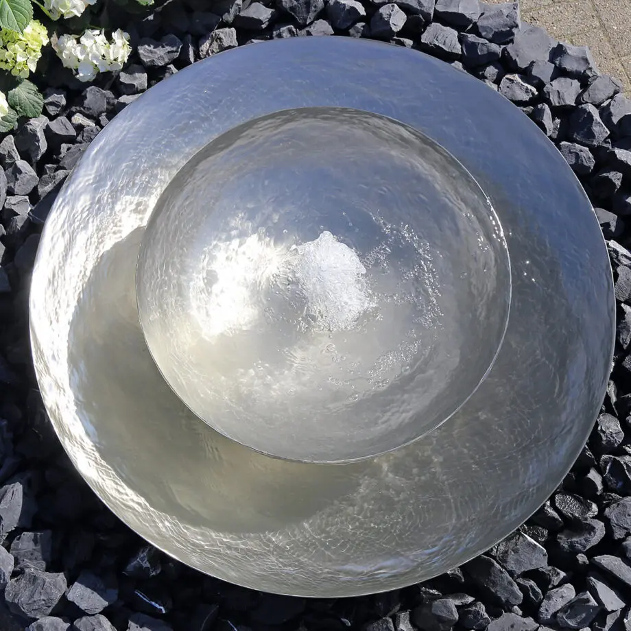 Edelstahl-Schale Duo mit zwei Wasserschalen (100 cm und 60 cm Ø), Sprudelquell in der oberen Schale, aufgestellt in einem modernen Garten mit Kies und Pflanzen.