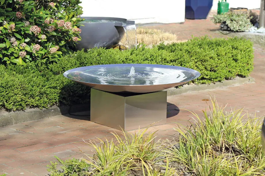 Edelstahl-Wasserschale Esbjerg mit 100 cm Durchmesser auf einem Edelstahlsockel, sanfte Wasserfontäne, aufgestellt in einem modernen Garten mit Kies und Pflanzen.
